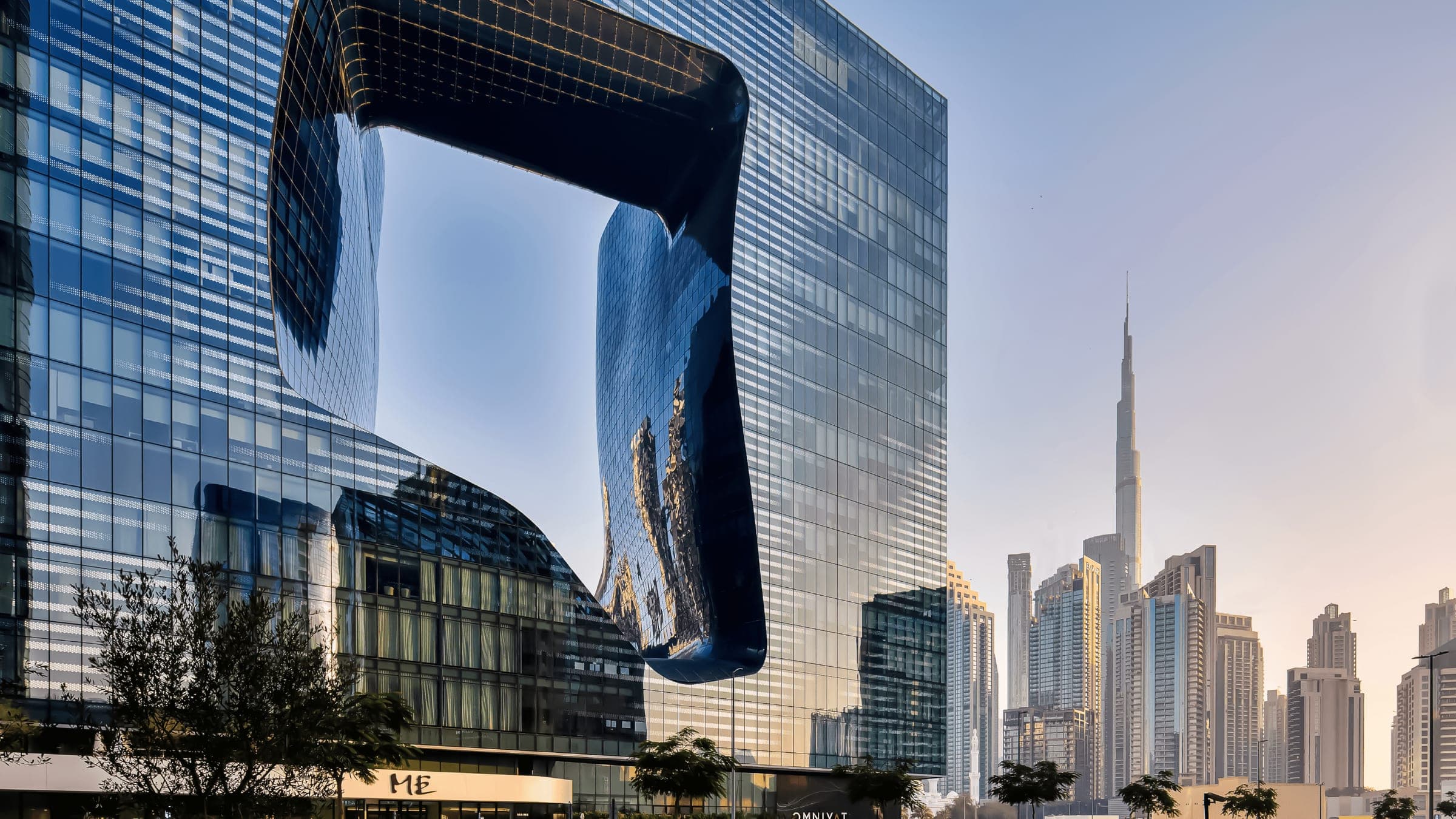 Photograph of The Opus by Omniyat in Business Bay — Zaha Hadid's glass cube with its free-form void — with the ME Dubai hotel sign at its base and Burj Khalifa behind